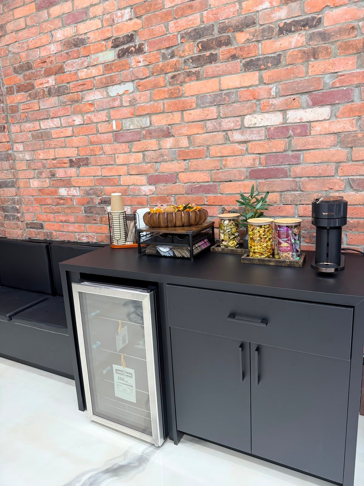 Complimentary coffee bar with an espresso machine, cups, snacks and a drinks fridge against a red brick wall at New Legend Barbershop in Hamilton